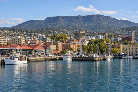 AI-generated editorial illustration of Hobart’s waterfront, with moored boats, city buildings and kunanyi / Mount Wellington behind.